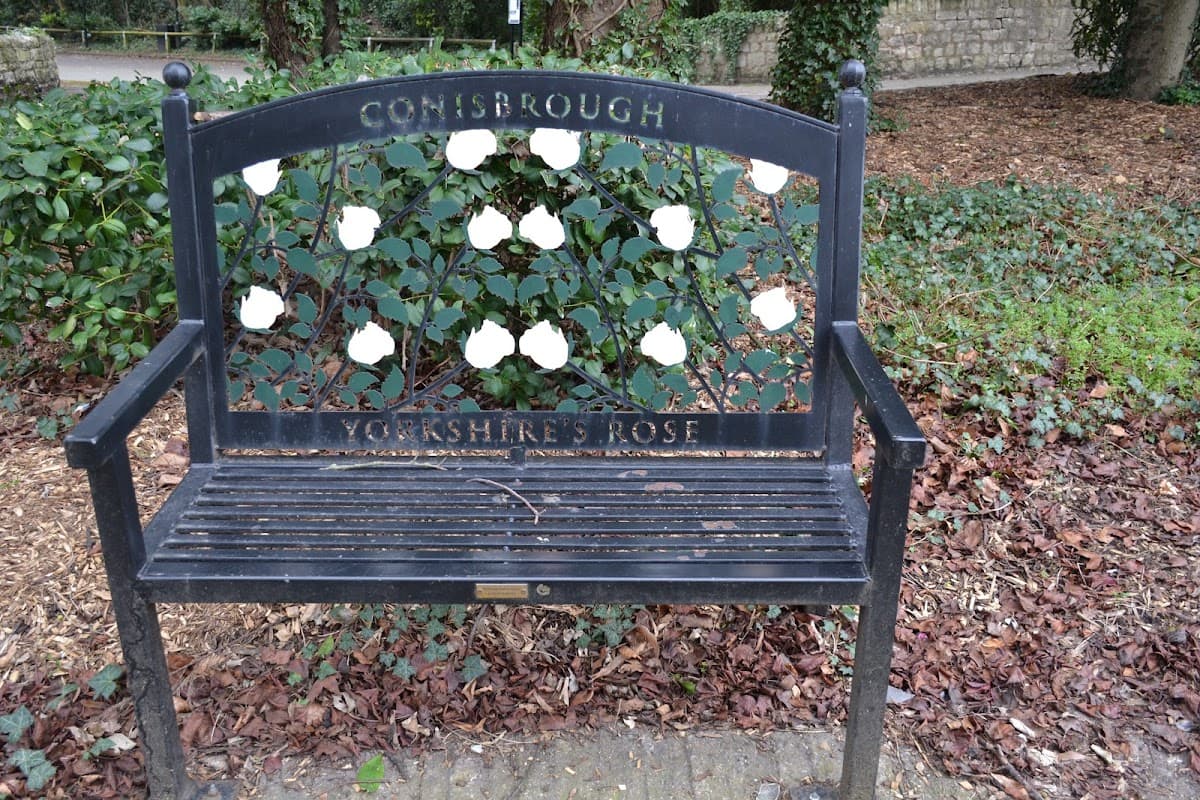 Black bench with white roses and "Conisbrough Yorkshire's Rose" engraved, surrounded by greenery and fallen leaves.