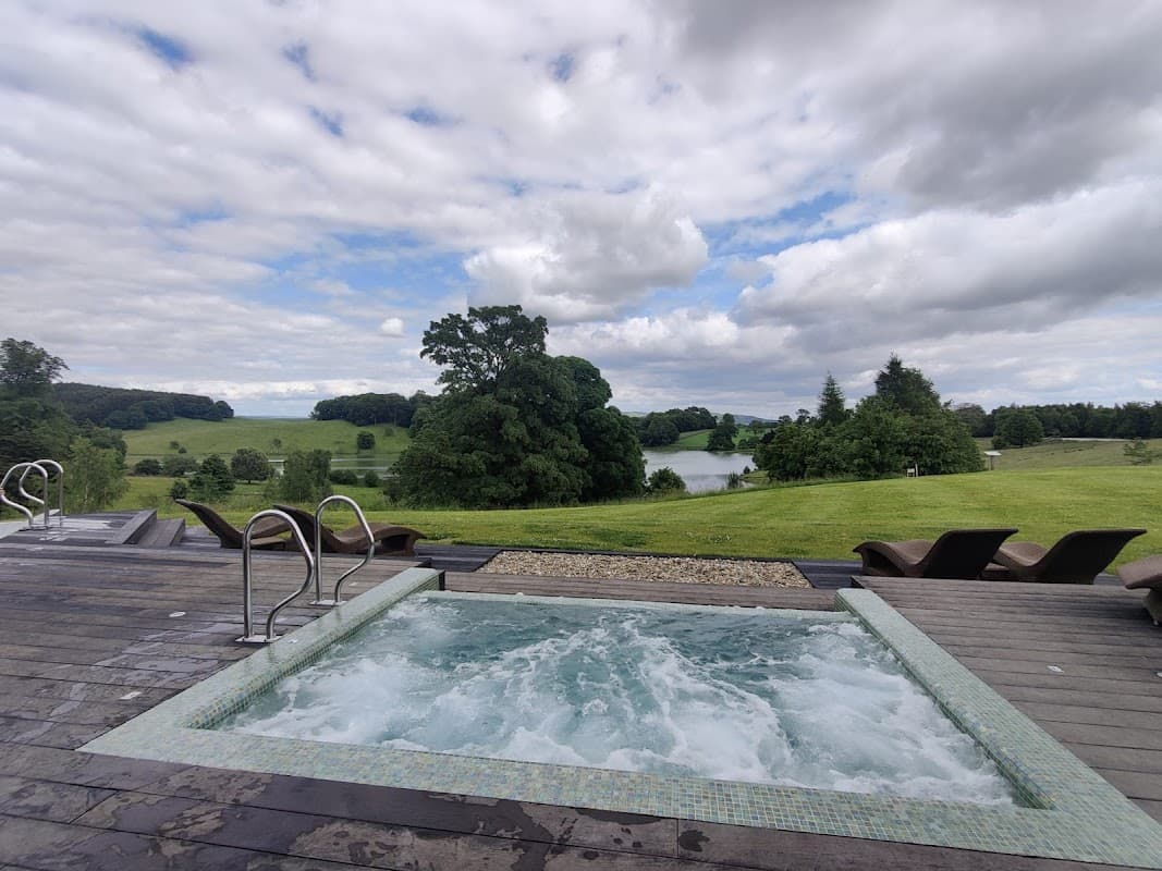 Hot tub on a wooden deck overlooking lush green fields and a tranquil lake under a cloudy sky.