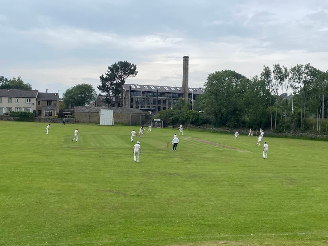 Cricket match in progress on a green field with buildings and trees in the background, under a cloudy sky.