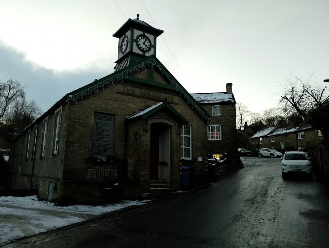 Cononley Village Institute features a clock tower, stone facade, and snowy surroundings with nearby houses on a sloped road.