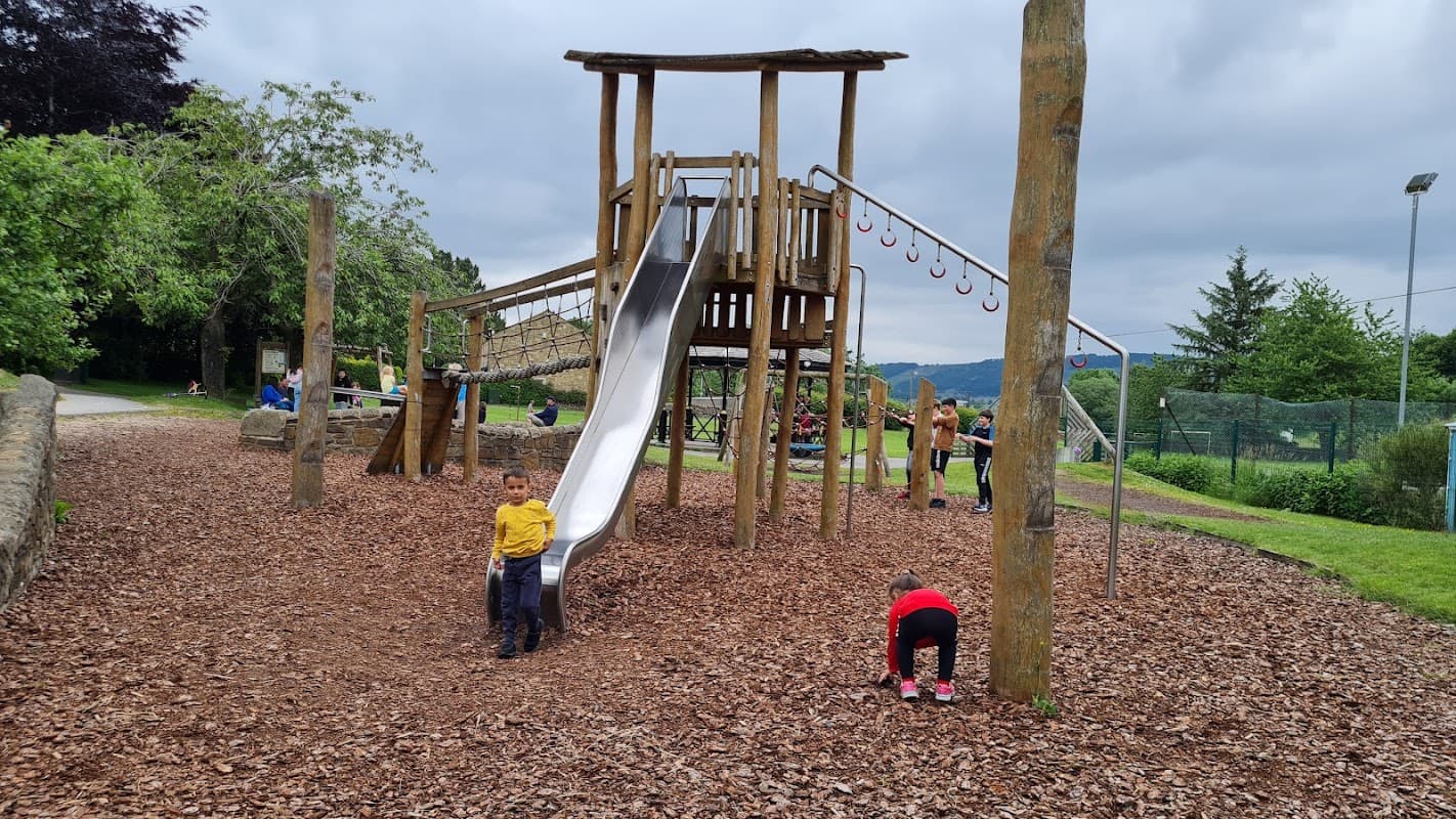 Wooden playground with a slide, climbing structure, and children playing on wood chips, surrounded by greenery.