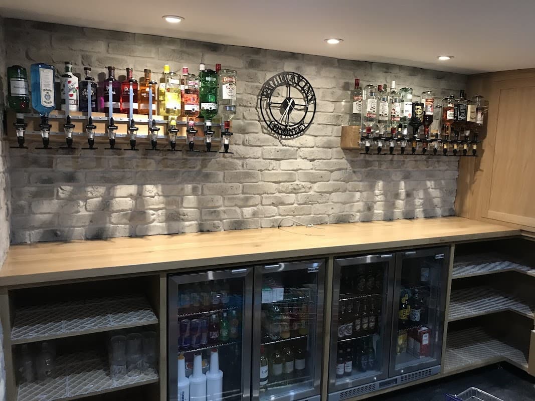 Bar area with a wooden counter, brick wall, and shelves displaying various bottles of spirits and a clock.