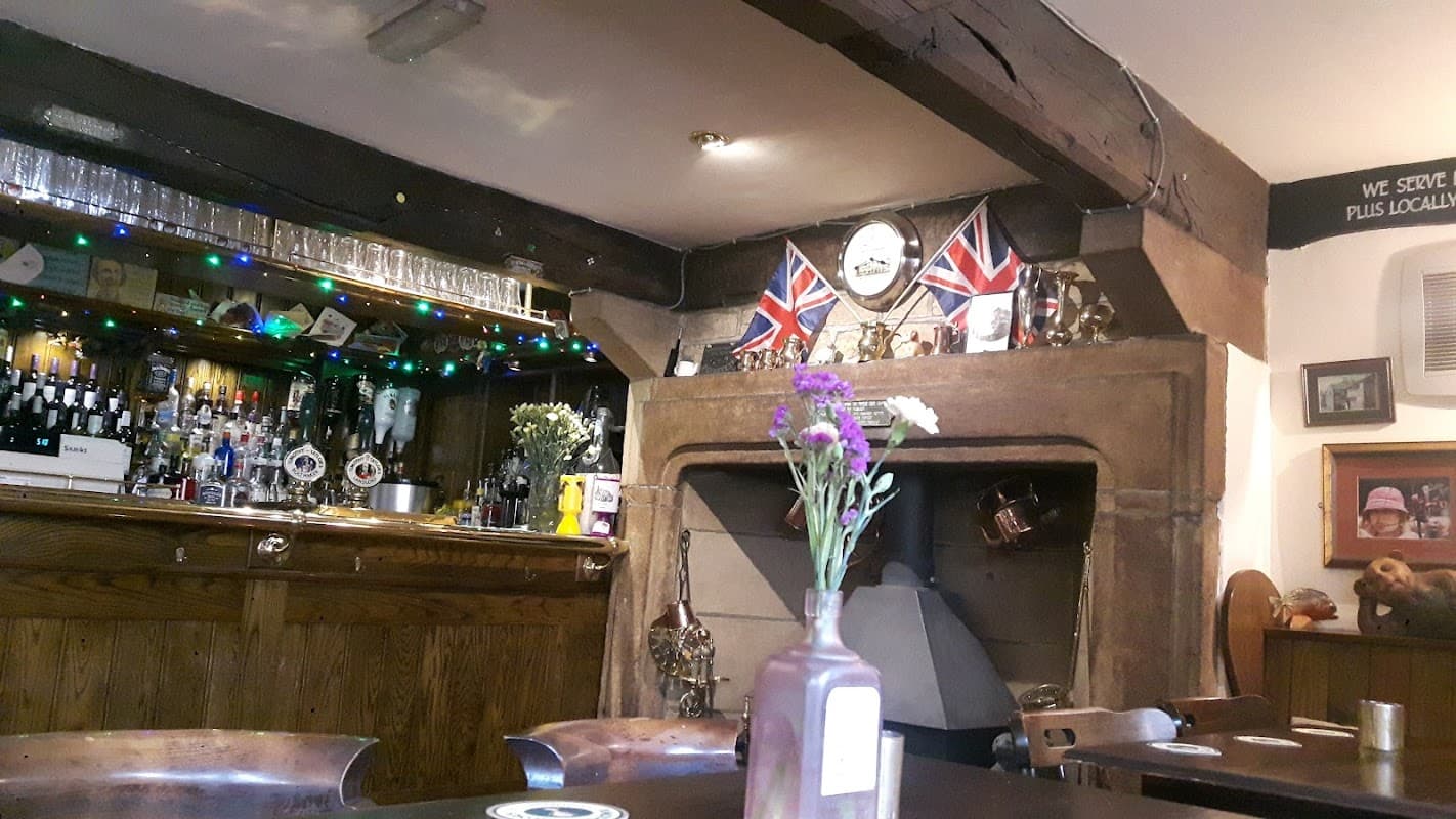 Cozy bar interior with wooden beams, a fireplace, a well-stocked bar, British flags, and a vase of flowers on the table.
