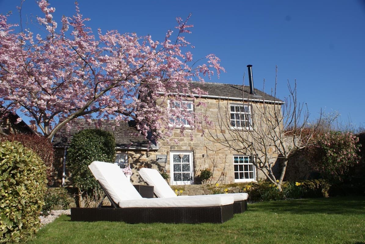 Charming stone cottage with blooming cherry trees and sun loungers in a lush garden, set against a clear blue sky.