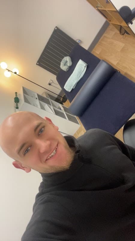 Bright wellness space with a treatment table, storage shelves, and a smiling individual in a black top.