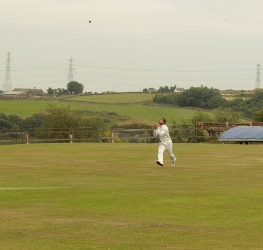A cricketer in white gear catches a ball on a grassy field with hills and power lines in the background.
