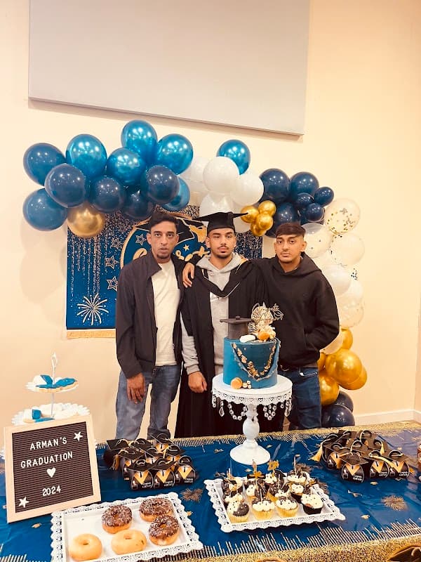 Graduation celebration with three young men posing by a decorated cake and dessert table, featuring blue and gold balloons.