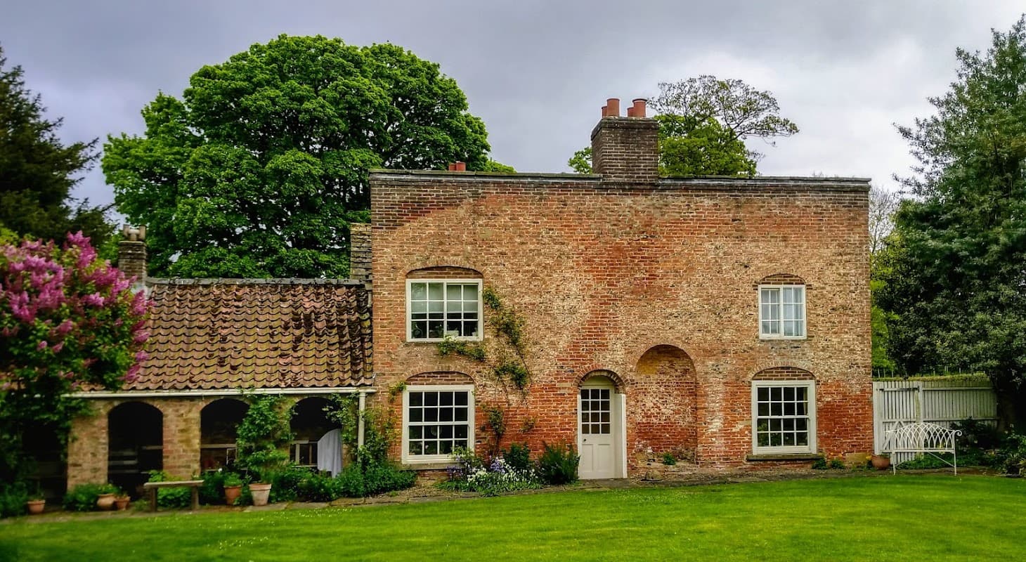A brick house with large windows, surrounded by lush greenery and flowering plants in a garden setting.