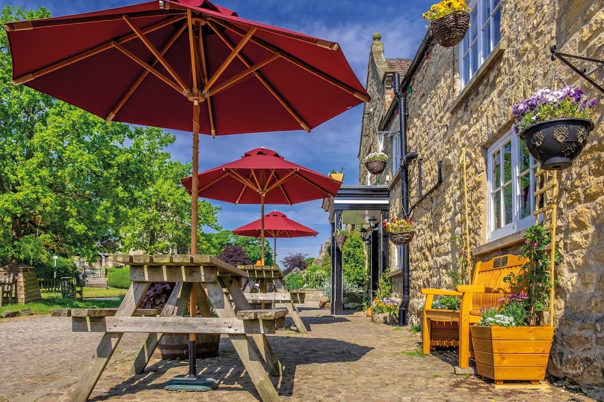 Stone building with red umbrellas over wooden tables, flowers in hanging baskets, and a sunny outdoor seating area.