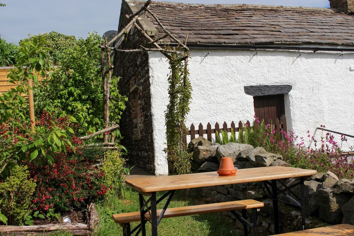 Charming white cottage with a stone wall, wooden door, and rustic garden tables surrounded by colorful flowers and greenery.