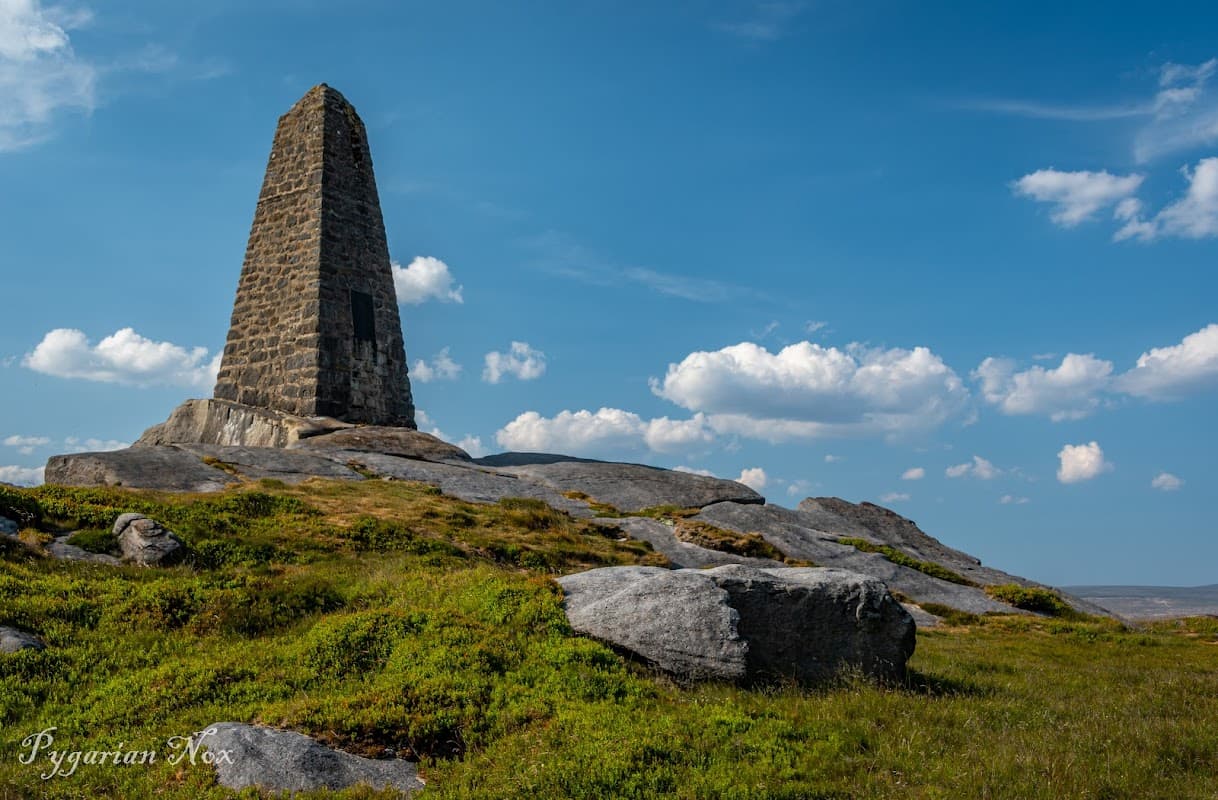 Cracoe War Memorial stands tall on rocky terrain under a blue sky with scattered clouds.