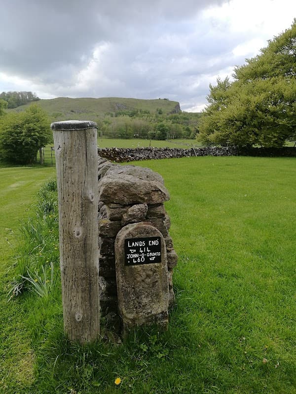 Signpost reading "Lands End" next to a stone pillar, with lush green grass and hills in the background.
