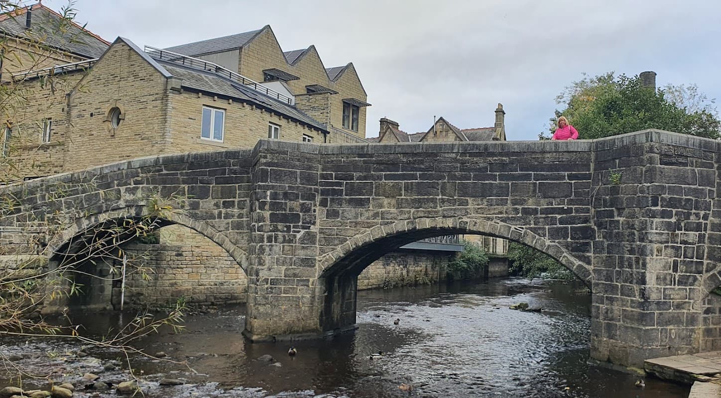 Stone bridge over a stream, with a person in a pink jacket standing on it, surrounded by historic buildings.