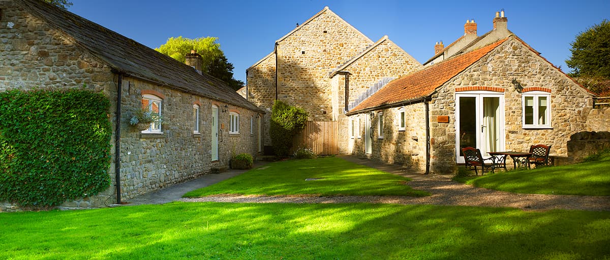 Stone buildings with slate roofs, a green lawn, and a small seating area under clear blue skies in Crakehall, Yorkshire.