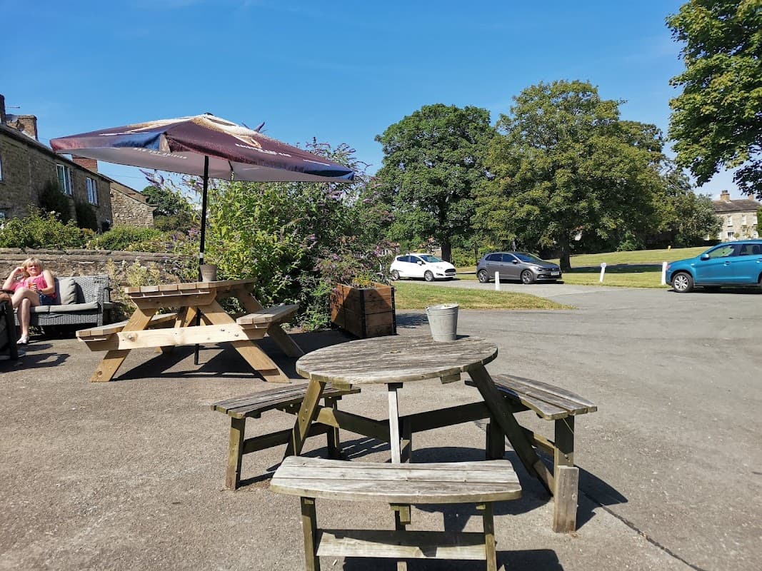 Outdoor seating area with wooden tables and umbrellas, surrounded by greenery and a clear blue sky in Crakehall, Yorkshire.
