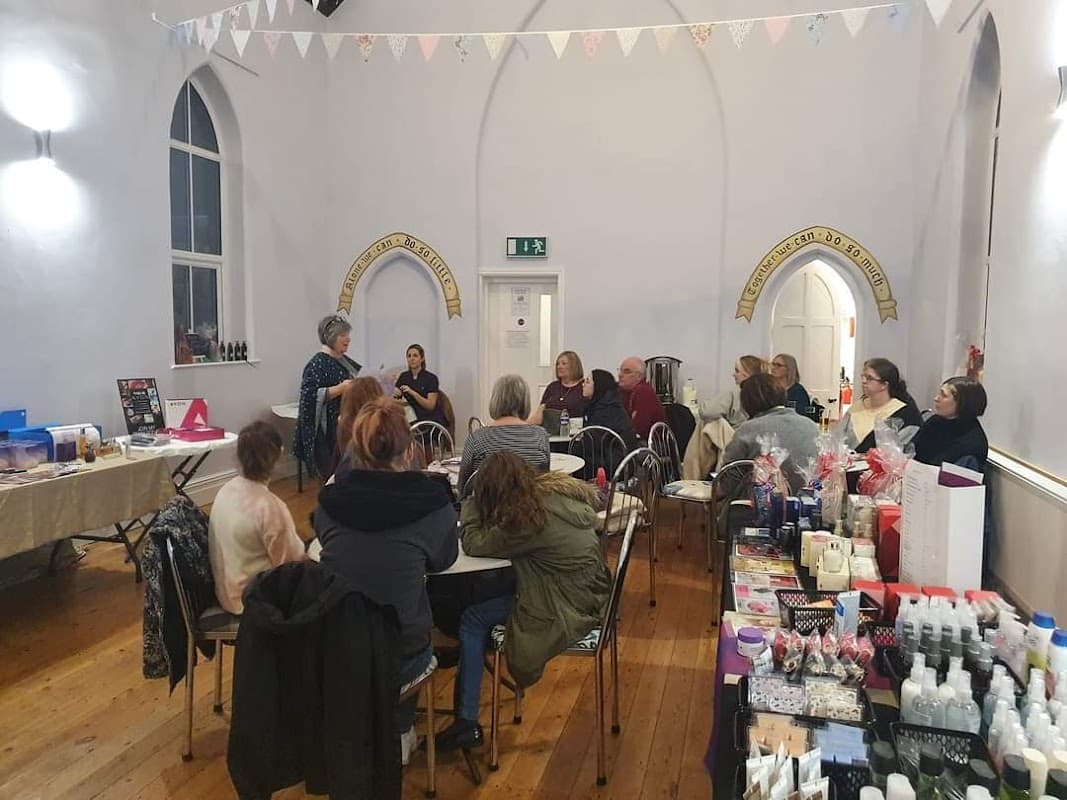 A group of people seated in a hall, listening to a speaker, with tables displaying various products and decorations.