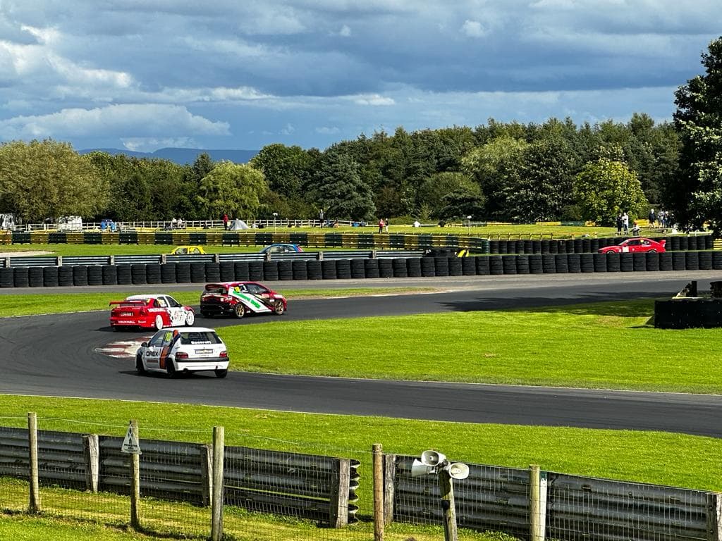 Cars racing on a track at Croft Circuit, surrounded by green trees and a cloudy sky.