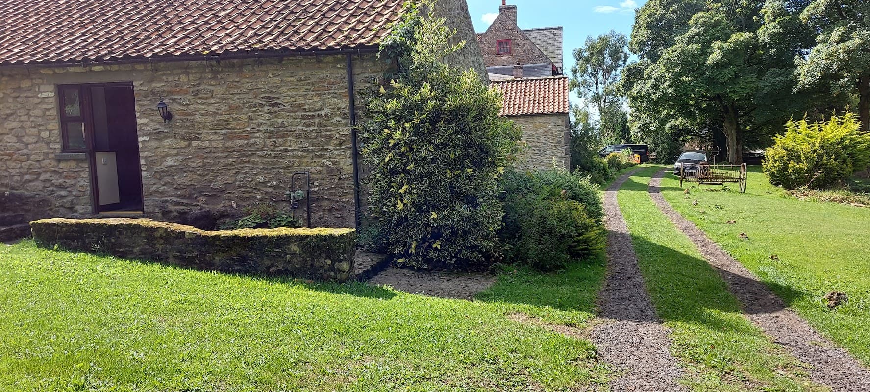 Stone cottage with a red-tiled roof, gravel path, lush greenery, and parked car in a tranquil Yorkshire setting.