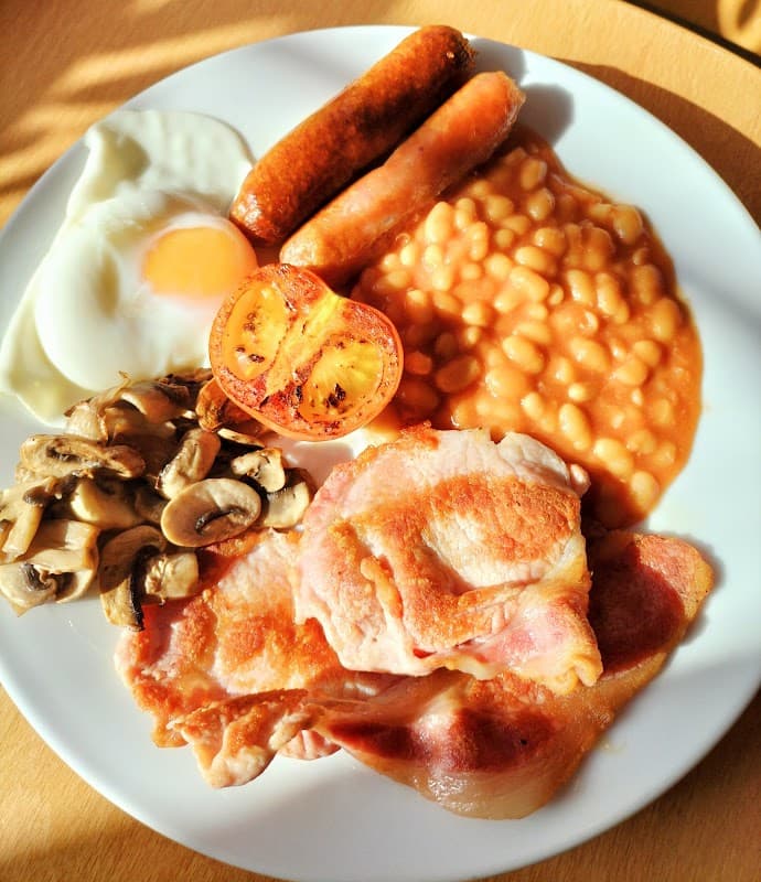 Full English breakfast featuring eggs, sausages, bacon, baked beans, mushrooms, and grilled tomatoes on a plate.