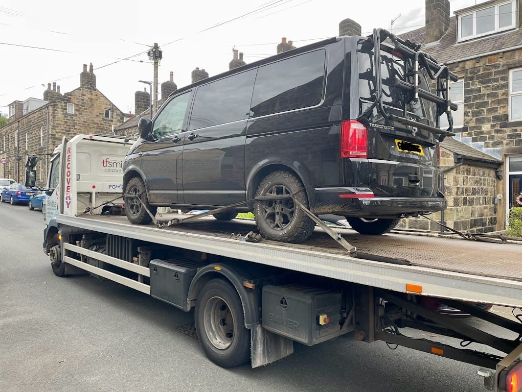 A black van on a recovery truck in a street lined with stone houses in Cross Hills, Yorkshire.