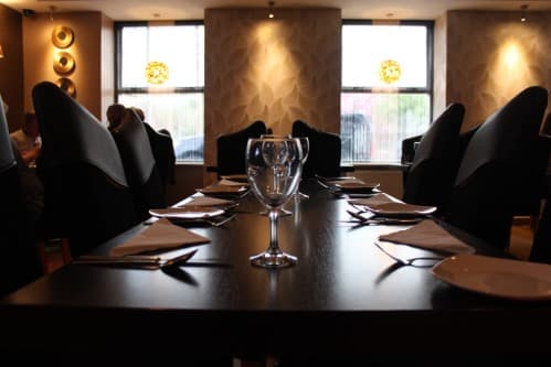 Elegant dining setup with a glass and cutlery on a dark table, featuring stylish chairs and soft lighting.