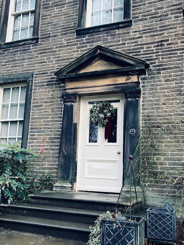 White door with a wreath, stone steps, and greenery beside the entrance of the Brontë Parsonage Museum.