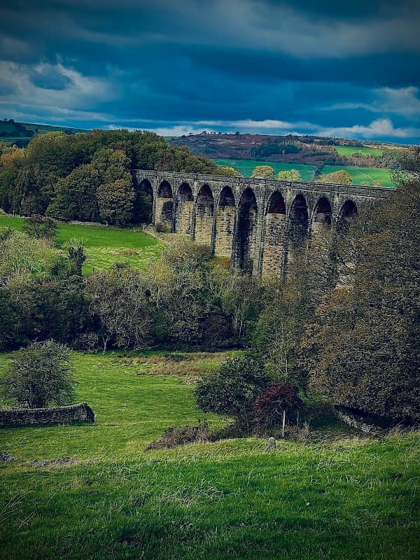 Hewenden Viaduct spans lush green fields, framed by trees under a dramatic cloudy sky in Cullingworth, Yorkshire.