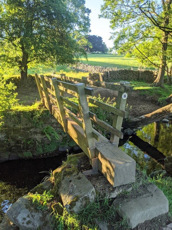 A wooden footbridge crosses a small stream, surrounded by lush greenery and trees in a tranquil campsite setting.