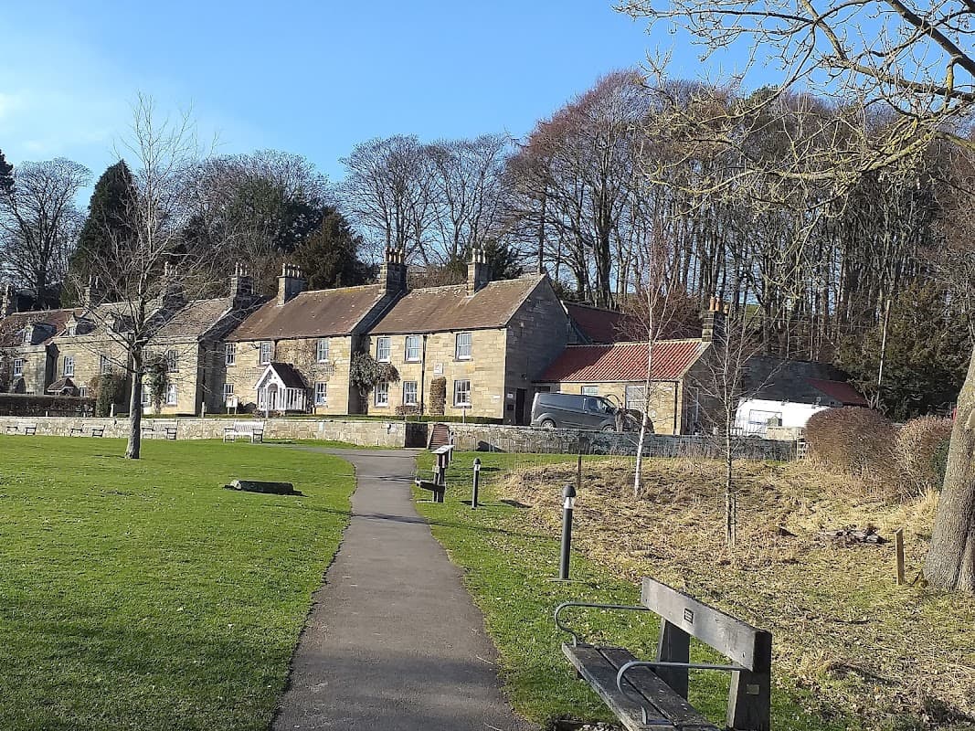 Pay & Display car park in Danby, featuring stone cottages, a pathway, benches, and surrounding trees.