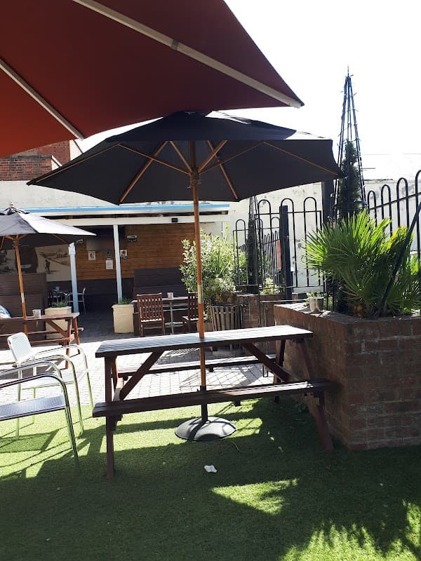 Outdoor seating area with a picnic table under a large umbrella, surrounded by greenery and a fence.