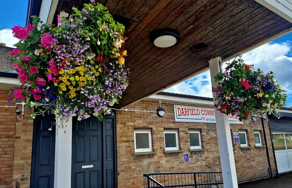 Colorful hanging flower baskets adorn the entrance of Darfield Community Centre, featuring a brick façade and signage.