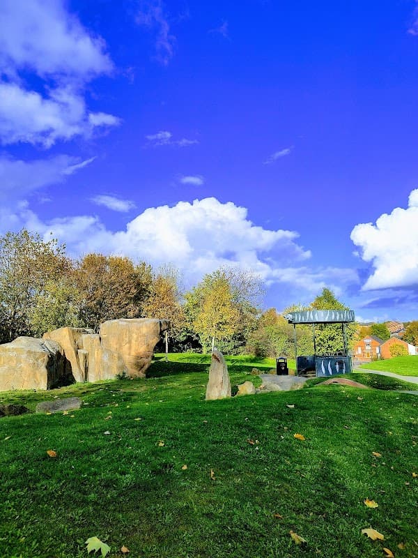 Lush green park with large rocks, trees, and a shelter under a bright blue sky with fluffy white clouds.