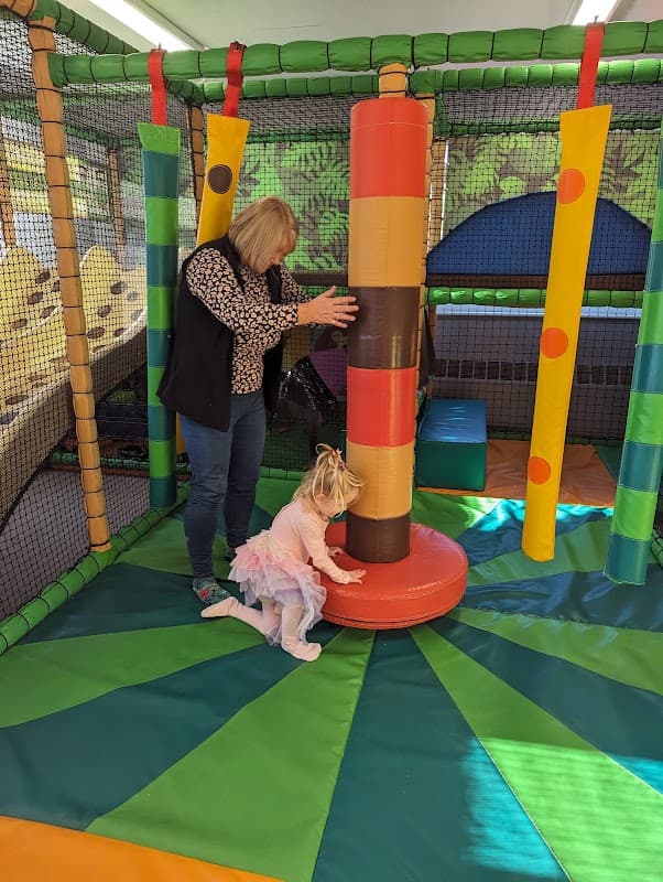 A child in a tutu plays with a colorful padded post while an adult assists in a vibrant indoor play area.