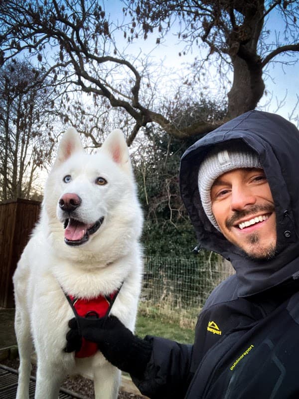 A man in a black jacket smiles beside a fluffy white dog with blue eyes in a park setting.
