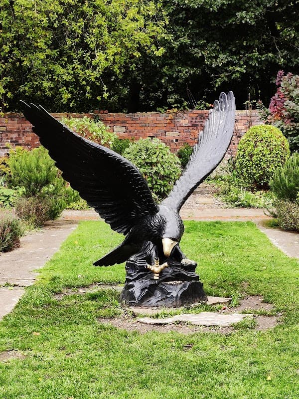 A large black eagle sculpture with outstretched wings perched on a rock, surrounded by greenery and a brick wall.
