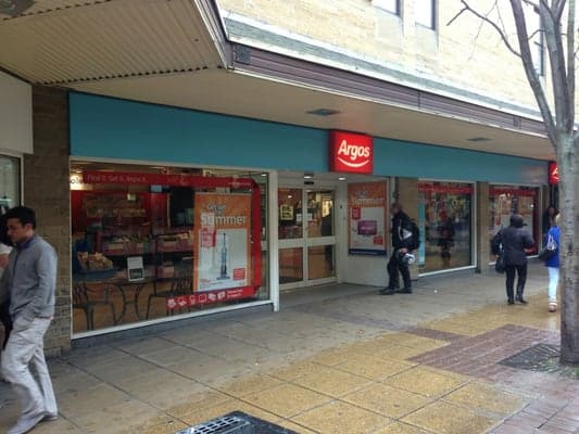 Argos storefront inside Sainsbury's, featuring large windows and promotional signage, with shoppers outside.