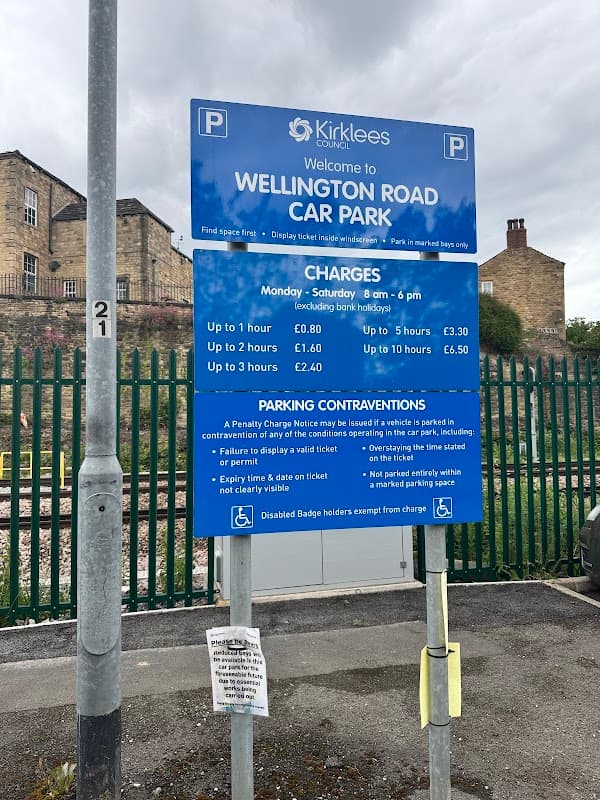 Sign for Wellington Road Car Park in Dewsbury, listing charges and parking regulations, with a cloudy sky above.