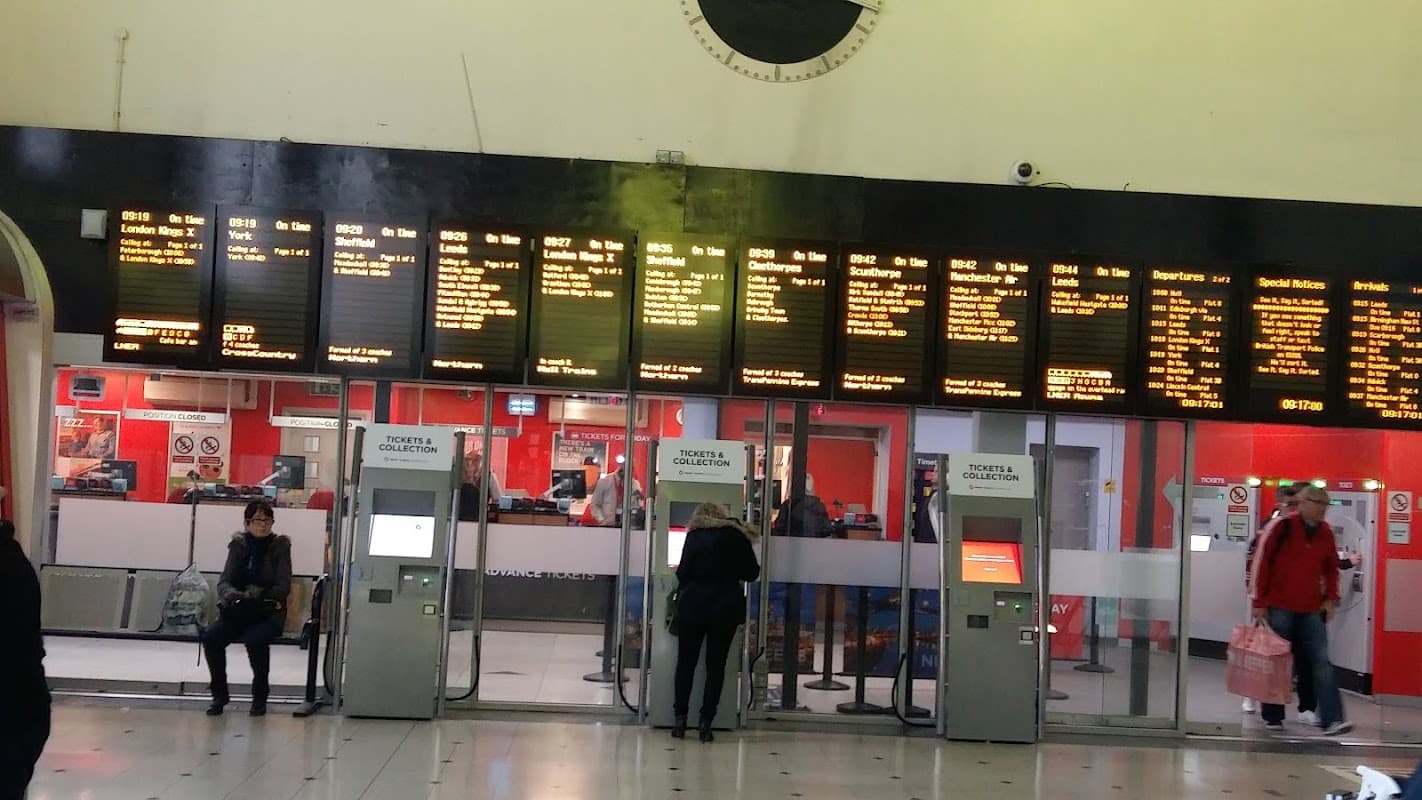 Digital display boards showing train times, ticket collection area, and passengers waiting in a busy station setting.