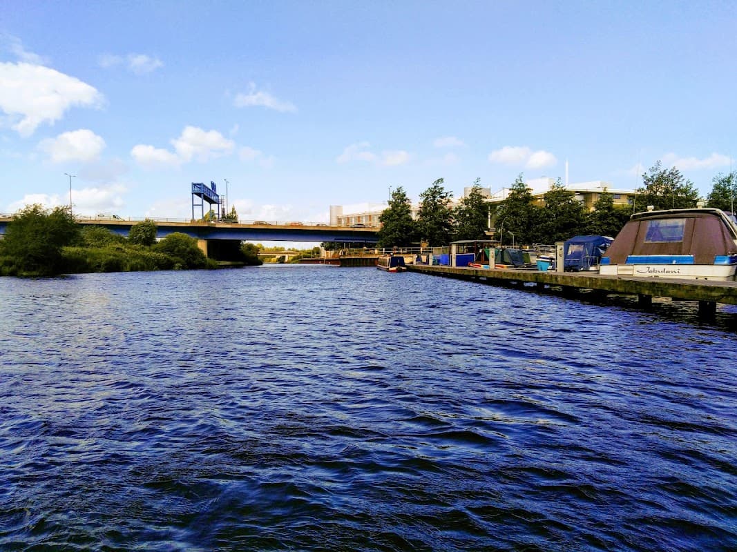 View of a river with boats and a car park in the background, under a clear blue sky with scattered clouds.
