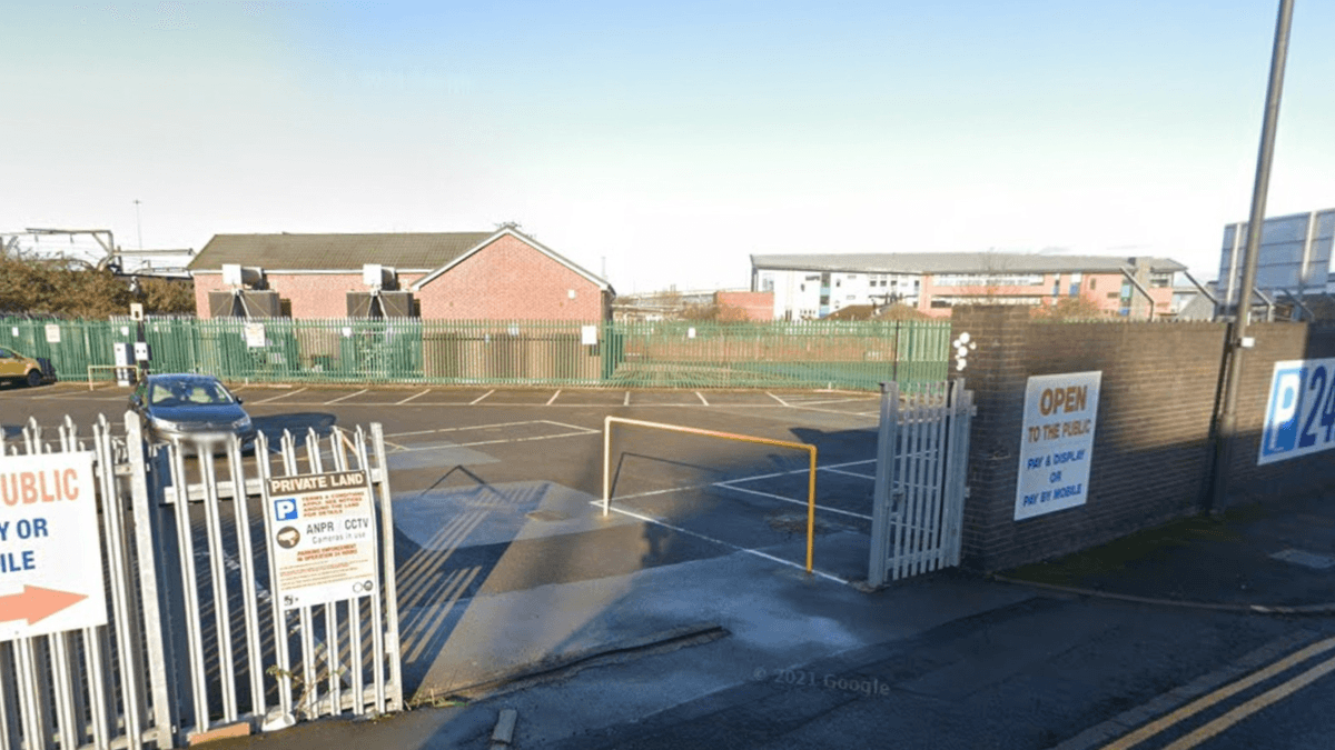 Parking lot entrance with gates, signage indicating open hours, and buildings in the background on Grey Friars Road.