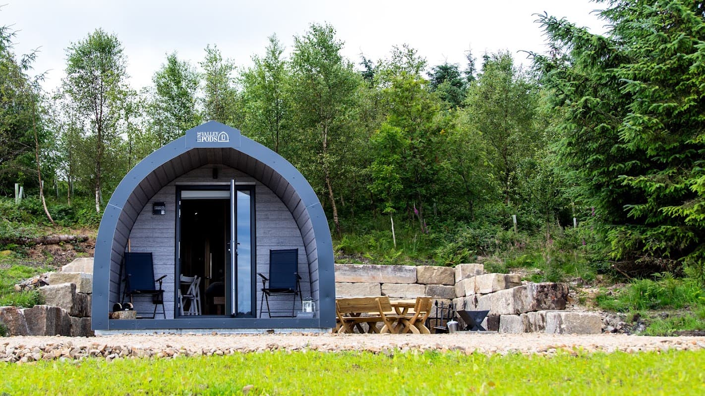 A cozy glamping pod with chairs on a stone patio, surrounded by lush greenery and trees at Draughton Heights Campsite.