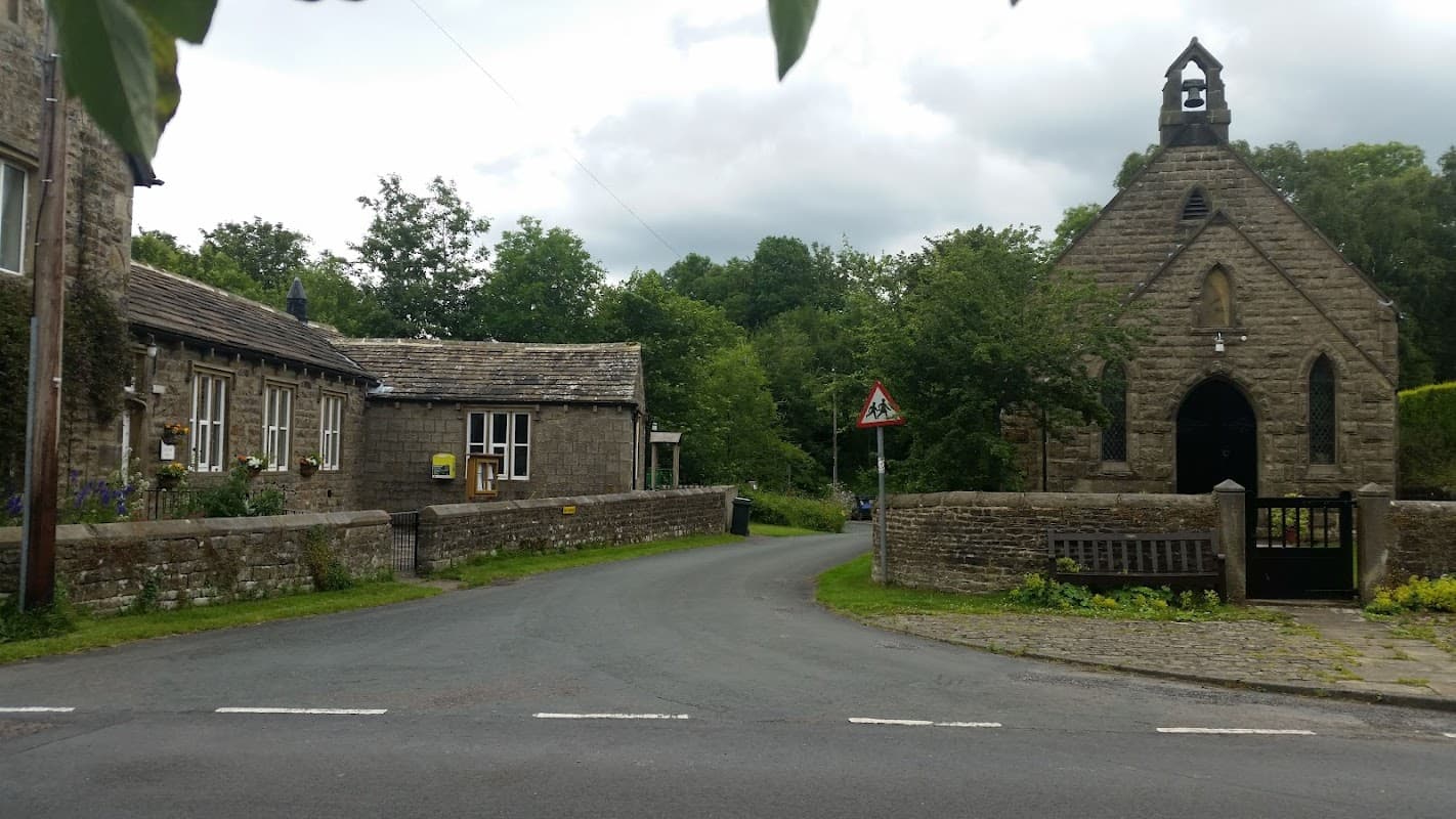 Draughton Village Hall with stone architecture, surrounded by greenery and a winding road, under a cloudy sky.