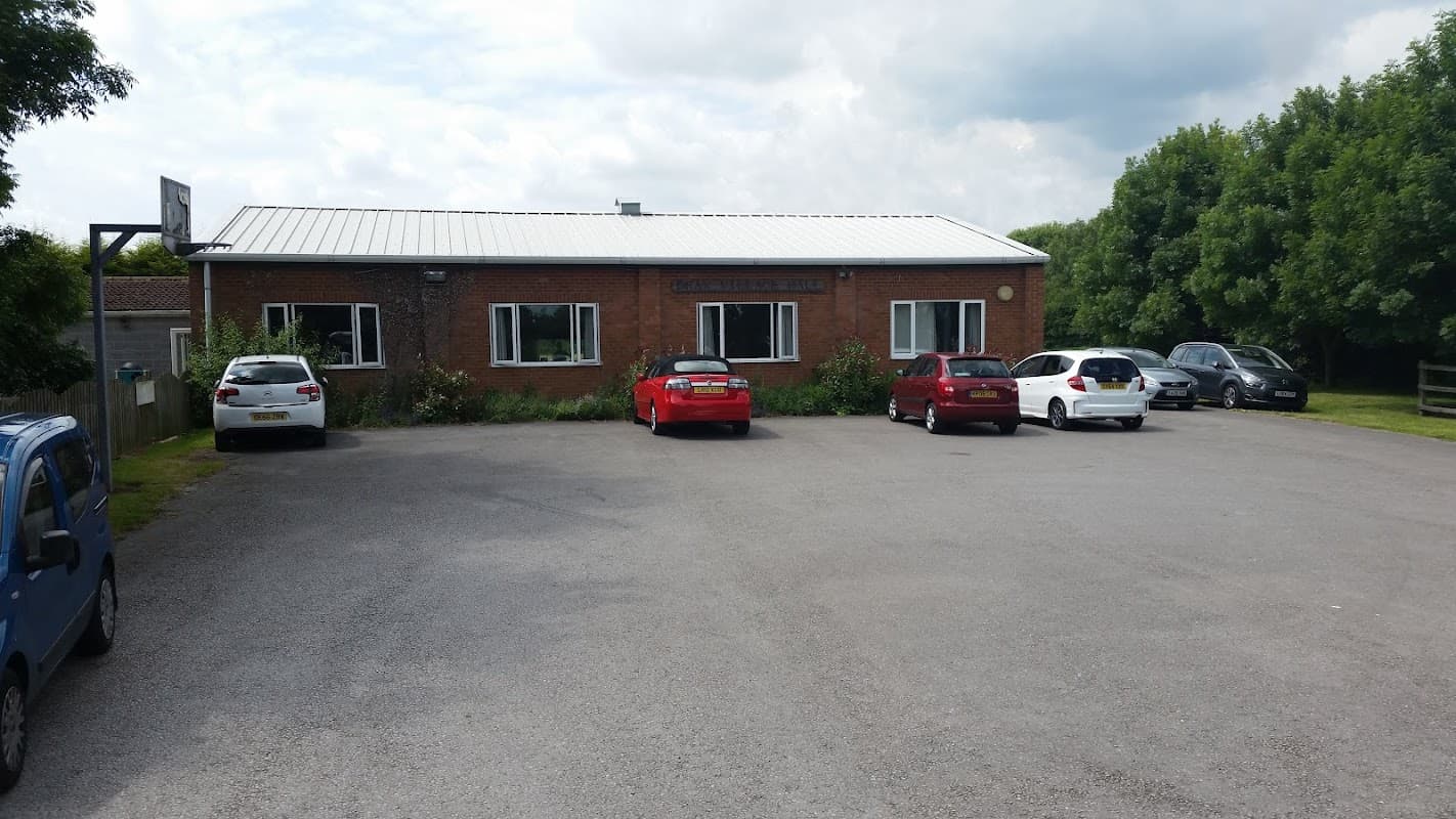 Drax Village Hall with a gravel parking lot and several parked cars, surrounded by greenery under a cloudy sky.