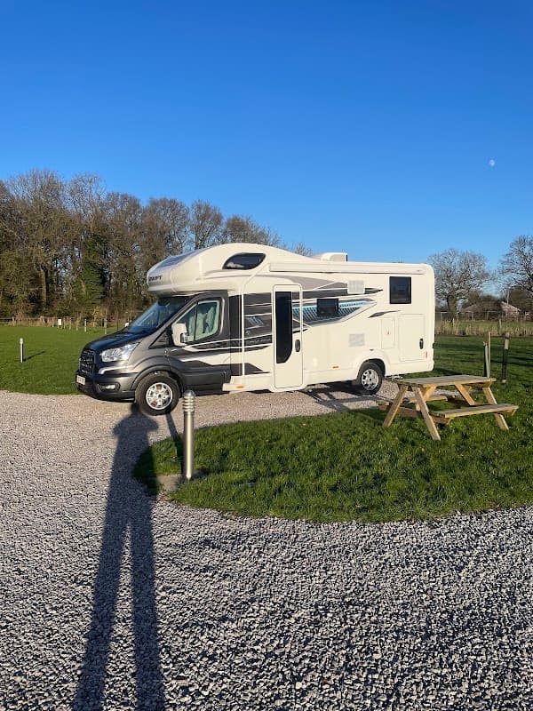 A camper van parked on gravel, surrounded by green grass and trees under a clear blue sky at Laurels Park Campsite.