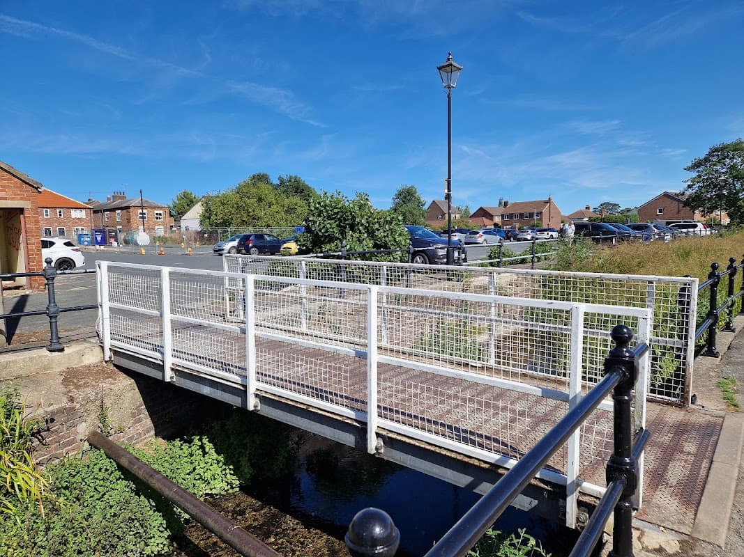 Bridge over a stream leading to Eastgate Free Car Park, with parked cars and grassy areas under a clear blue sky.
