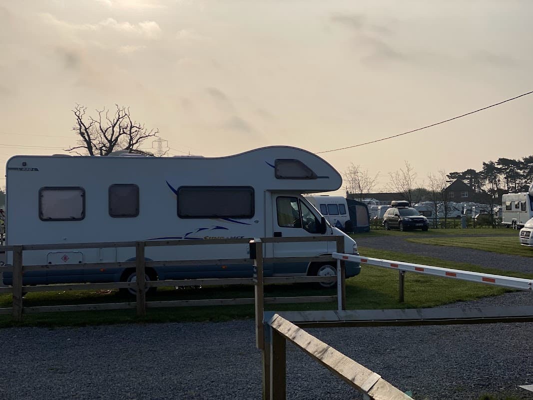 Caravans parked on a grassy site, with trees and a cloudy sky in the background.