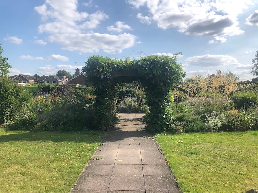 A lush garden path leads through a green archway, surrounded by colorful flowers and under a bright blue sky.