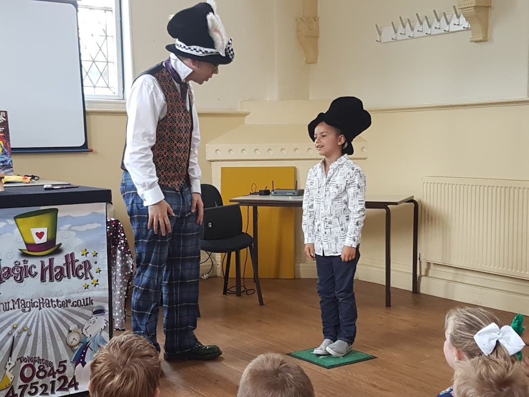 A magician performs for a child wearing a top hat, with an audience of children seated in the background.