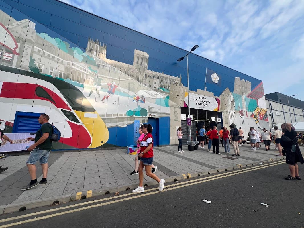Crowd of people walking past a colorful mural and entrance to York Community Stadium, with parked cars nearby.