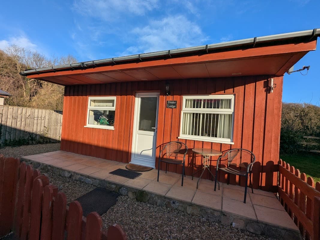 Red wooden chalet with a white door, two chairs outside, and a gravel pathway in a scenic outdoor setting.